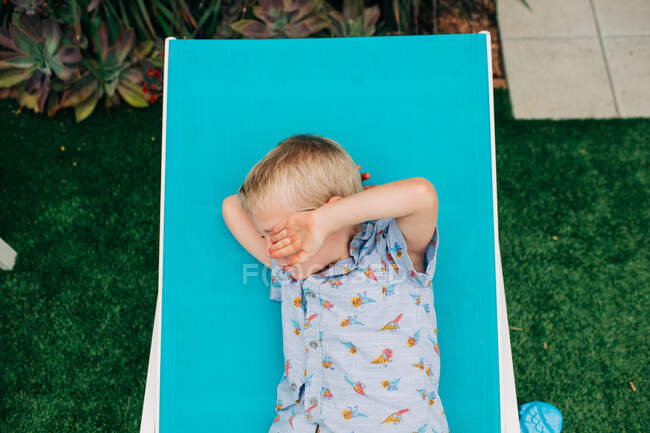 Menino loiro protege os olhos do sol enquanto descansa na cadeira — Fotografia de Stock