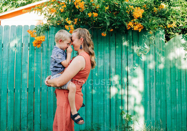 Madre acurruca a niño pequeño mientras está de pie frente a la cerca verde - foto de stock