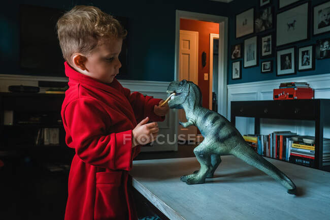 Toddler in red bathrobe brushing a t-rex's teeth — Stock Photo