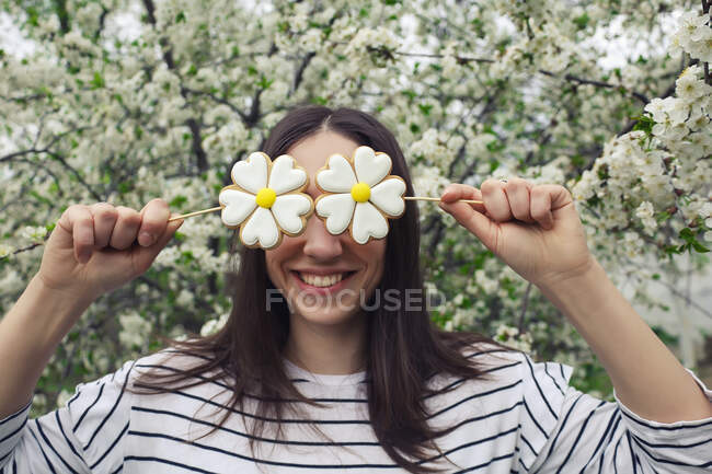 Усміхнена дівчина приховує очі з імбирним тістечком на паличках у ромашковій квітковій формі — стокове фото