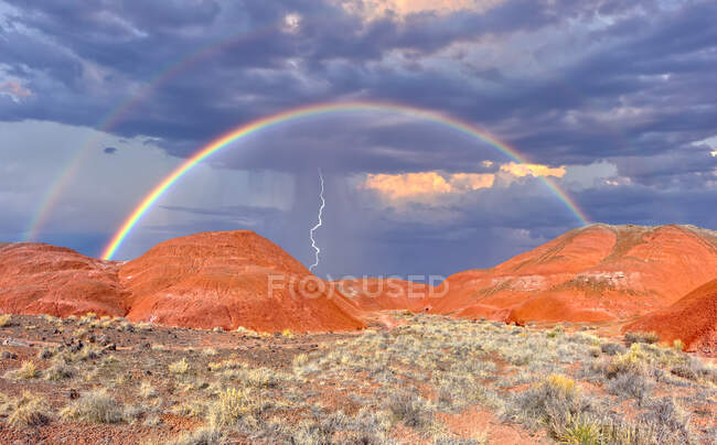 Un HDR compuesto de un arco iris y una tormenta acercándose a las colinas de bentonita roja debajo de Kachina Point del Parque Nacional Bosque Petrificado de Arizona. El paisaje y el arco iris fueron capturados antes de que la lluvia llegara a esta área específica. El rayo fue capturado - foto de stock