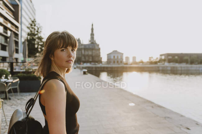 Young woman with long hair in a white dress and sunglasses sitting on the bridge at the river bank — Stock Photo