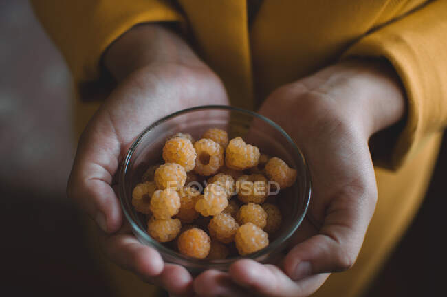 Yellow raspberries in a glass plate in the hands of a girl in a yellow jacket — Stock Photo