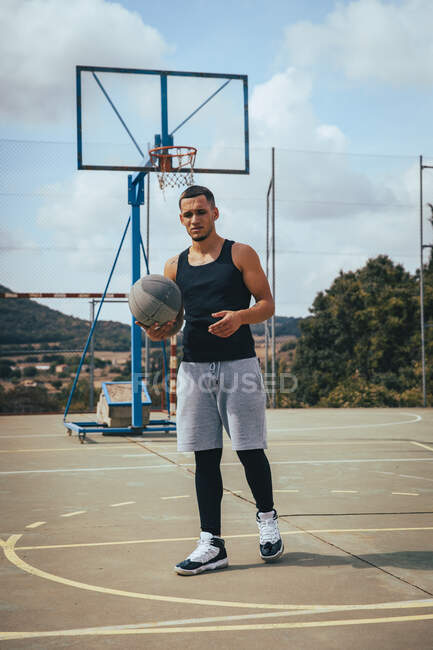 Joven chico latino tatuado jugando con una pelota de baloncesto en una cancha - foto de stock