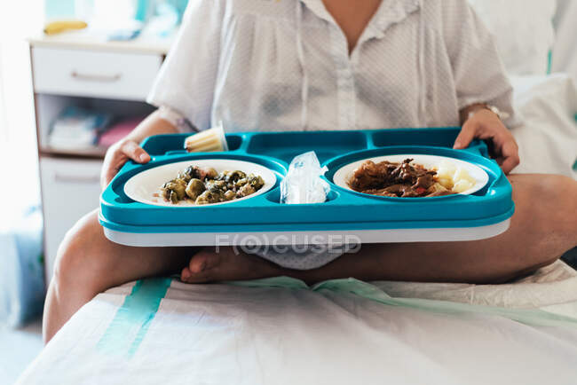 Mujer joven hospitalizada en una cama. Sostiene bandeja de comida hospitalaria. - foto de stock