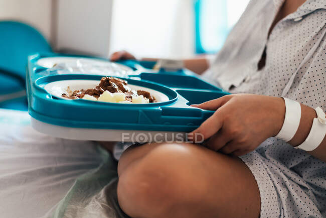 Jovem hospitalizada em uma cama. A segurar bandeja de comida do hospital. Vista lateral. — Fotografia de Stock