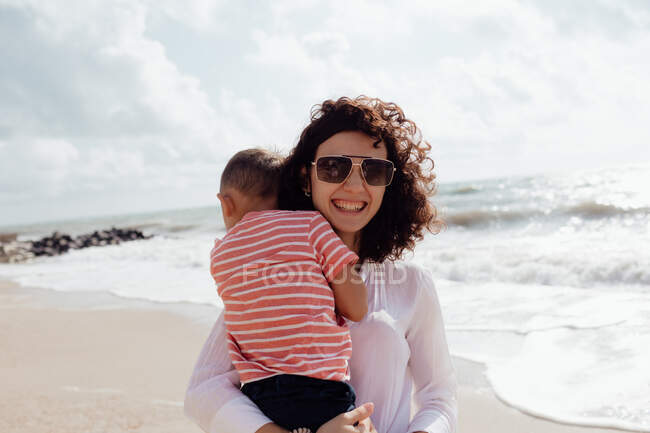 Joven hermosa mamá jugando con su hijo bebé en la playa - foto de stock