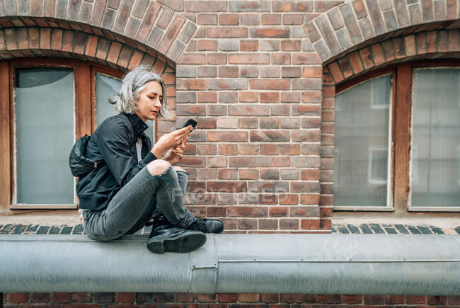 A woman sits cross-legged against the background of a brick building and looks at the smartphone screen — Stock Photo