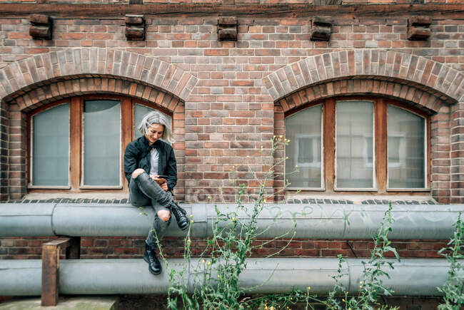 Uma mulher senta-se em um tubo perto de um edifício abandonado, olha para a tela do smartphone e sorri — Fotografia de Stock