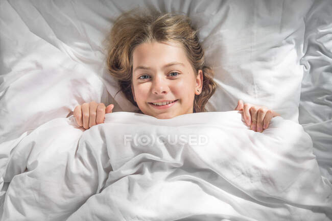 La chica mira desde debajo de las sábanas en una mañana soleada - foto de stock