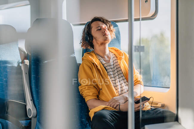 Молодой человек, подросток, путешествующий в поезде в наушниках, слушающий музыку. Снимок образа жизни с копированием пространства. — стоковое фото