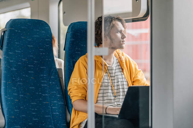 Jeune homme, adolescent, voyageant en train, regardant par la fenêtre, travaillant sur un ordinateur portable. Mode de vie tourné avec espace de copie. — Photo de stock