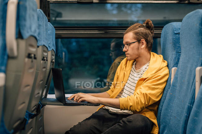 Young teenage man with glasses travels in train with laptop, programmer works remotely. Horizontal shot, portrait of traveler. — Stock Photo