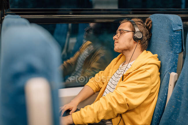 Young teenager in headphones listen to music, close eyes, travels in train. Shot with copy space and reflection in window. — Stock Photo