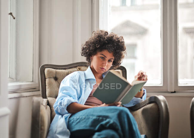 Молода жінка читає книгу, сидячи на кріслі — стокове фото
