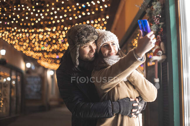 Pareja romántica a pie y tomar una videollamada a familiares en la ciudad con decoraciones navideñas - foto de stock