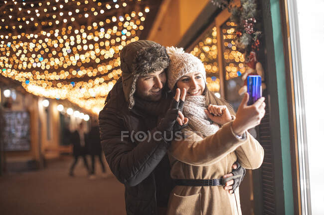 Pareja romántica a pie y tomar una videollamada a familiares en la ciudad con decoraciones navideñas - foto de stock