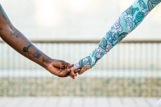 Crop unrecognizable best diverse female friends with tattoos in ornamental wear holding hands in town on blurred background — Stock Photo