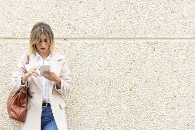 Mujer de negocios joven y moderna apoyada en una pared mirando al móvil - foto de stock