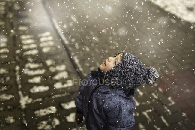 Ragazzino in abiti invernali blu guardando il cielo nevicare — Foto stock