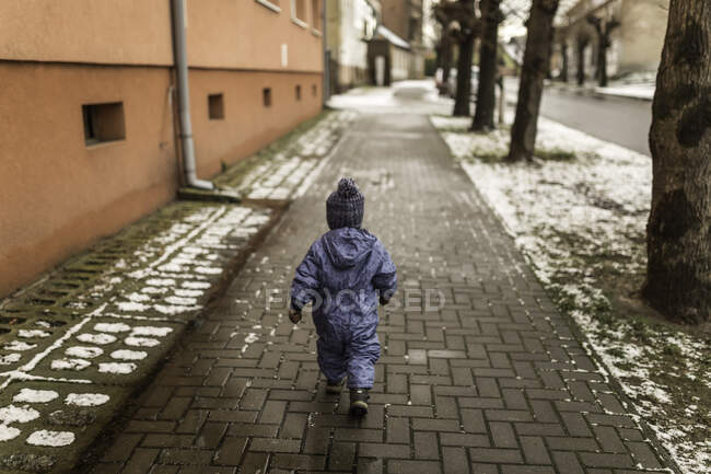Little boy in blue onesie and winter hat walking on pavement — Stock Photo