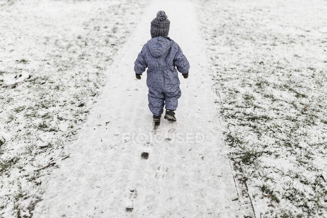 Маленький мальчик в голубых ползунках и теплая шляпа, идущий по снегу — стоковое фото
