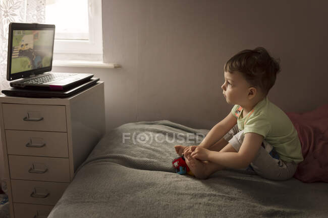 Tout-petit garçon assis sur le lit et regardant dessin animé sur ordinateur portable — Photo de stock