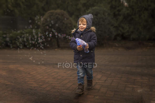 Toddler in warm clothes playing with bubble gun outside — Stock Photo
