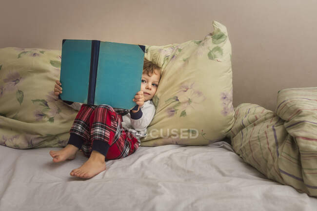 Toddler in pajamas sitting on bed and reading bedtime book looki — Stock Photo