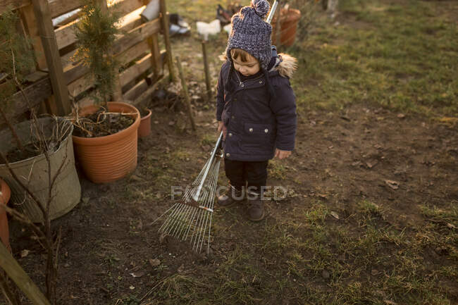 Kleiner Junge in Winterkleidung harkt Boden mit großem und rostigem Ra — Stockfoto