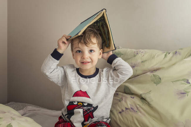 Niño en pijama poniéndose un libro en la cabeza y sentado en una cama - foto de stock