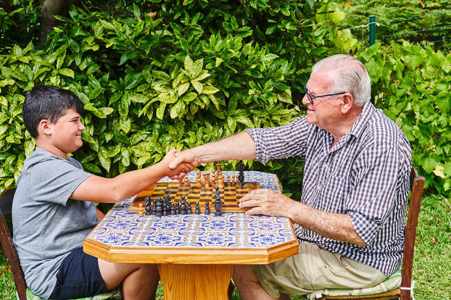 Avô e neto apertando as mãos sobre o tabuleiro de xadrez — Fotografia de Stock