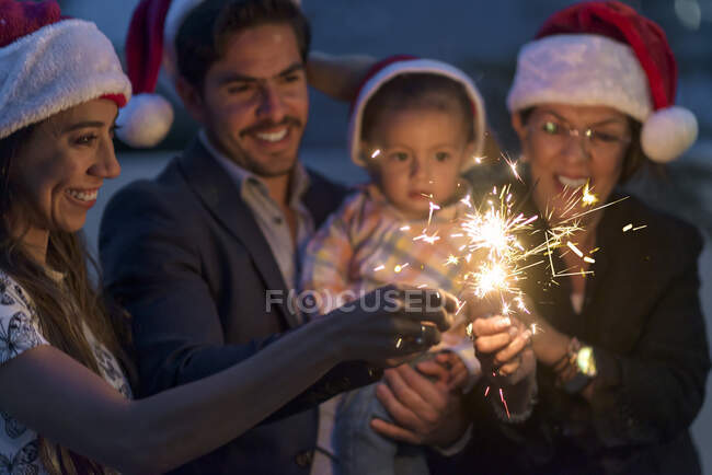 Família latina comemorando feriados de Natal ano novo se divertindo com faíscas ao amanhecer vestindo chapéus de Papai Noel — Fotografia de Stock