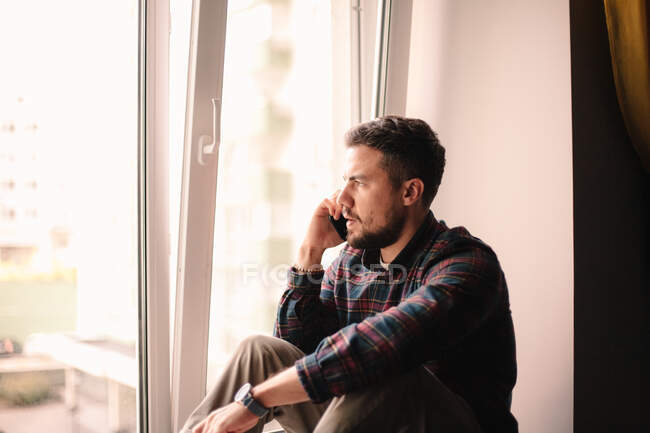 Серьезный человек разговаривает по смартфону сидя у окна дома — стоковое фото