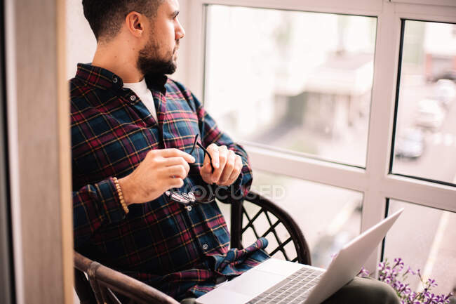 Homem pensativo olhando através da janela trabalhando com laptop em casa — Fotografia de Stock