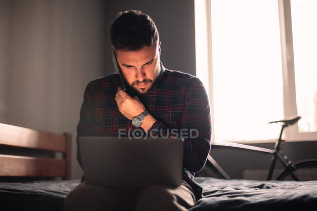 Man talking on smart phone using laptop computer working at home — Stock Photo