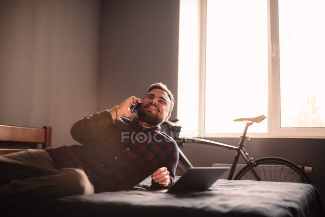 Homem feliz falando no telefone inteligente deitado na cama em casa — Fotografia de Stock