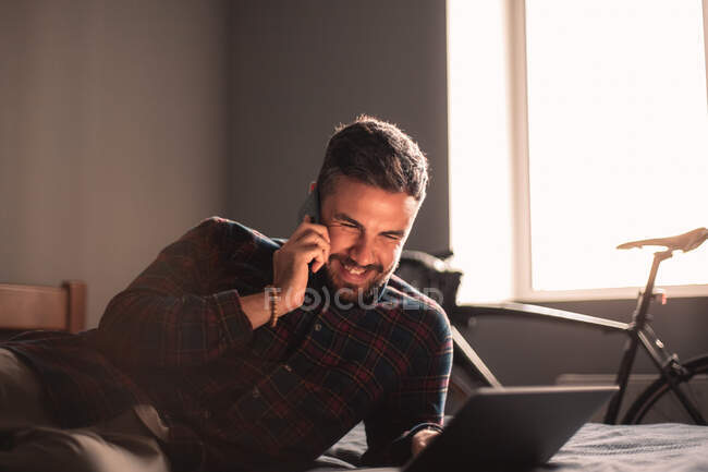 Glücklicher Geschäftsmann, der zu Hause mit dem Laptop auf dem Smartphone spricht — Stockfoto