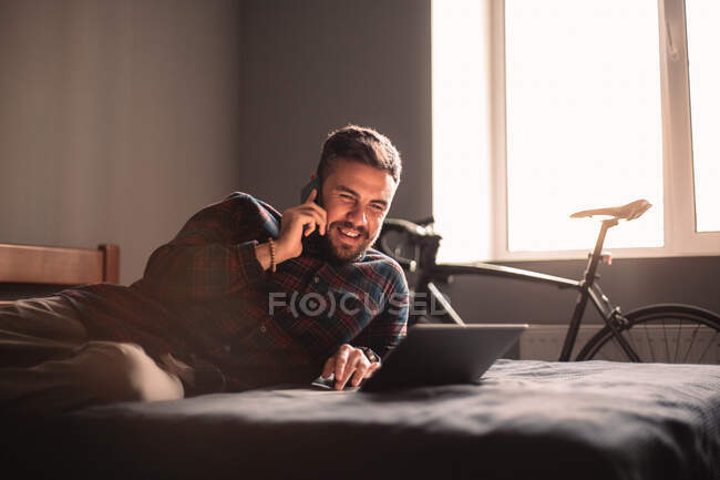 Щасливий чоловік розмовляє на смартфоні, використовуючи ноутбук вдома — стокове фото