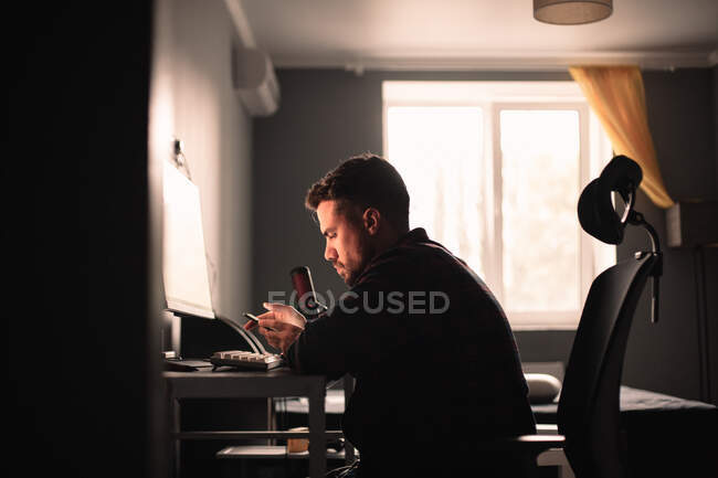 Чоловік, використовуючи смартфон і комп'ютер, сидить за столом, працює вдома — стокове фото