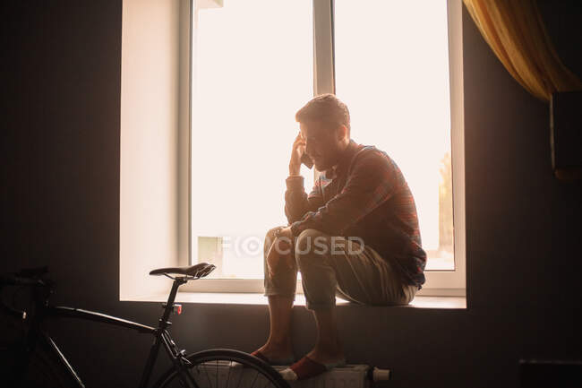 Hombre hablando por teléfono inteligente sentado en el alféizar de la ventana en casa - foto de stock