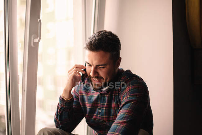 Щасливий чоловік розмовляє по смартфону, сидячи біля вікна вдома — стокове фото