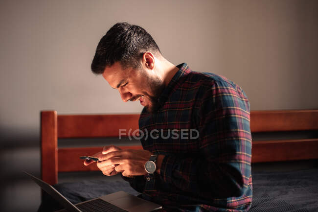 Homme heureux utilisant téléphone intelligent et ordinateur portable assis à la maison — Photo de stock
