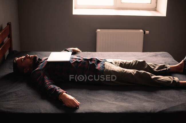 Uomo esausto con computer portatile sul petto sdraiato sul letto a casa — Foto stock