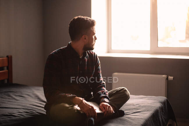 Hombre reflexivo con el ordenador portátil mirando a través de la ventana sentado en la cama en casa - foto de stock