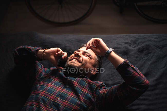 Веселый мужчина разговаривает по смартфону лежа на кровати дома — стоковое фото