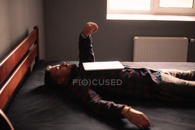 Müder Mann schaut auf Uhr, die zu Hause auf dem Bett liegt, Laptop auf dem Kopf — Stockfoto