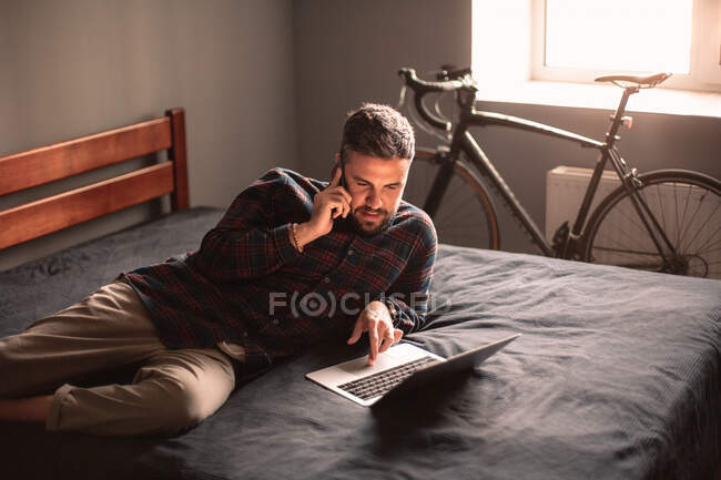 Мужчина разговаривает на смартфоне, используя ноутбук, работающий дома — стоковое фото