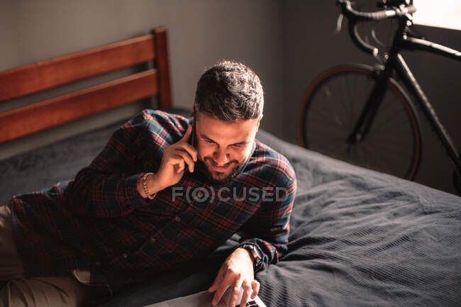 Homem feliz falando no telefone inteligente ao usar o computador portátil em casa — Fotografia de Stock