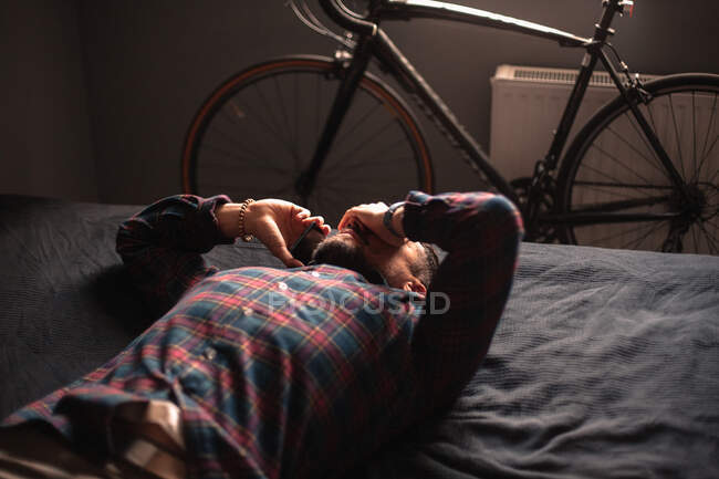 Виснажений чоловік розмовляє на смартфоні, лежачи в ліжку вдома — стокове фото